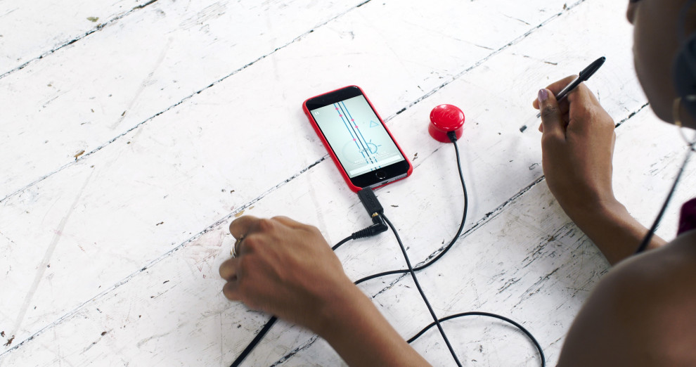 Eine Person sitzt an einem Tisch, auf dem ein Smartphone liegt. Das Telefon hat Kopfhörer angeschlossen und ein Kabel, das zu einem roten Knopf führt. 