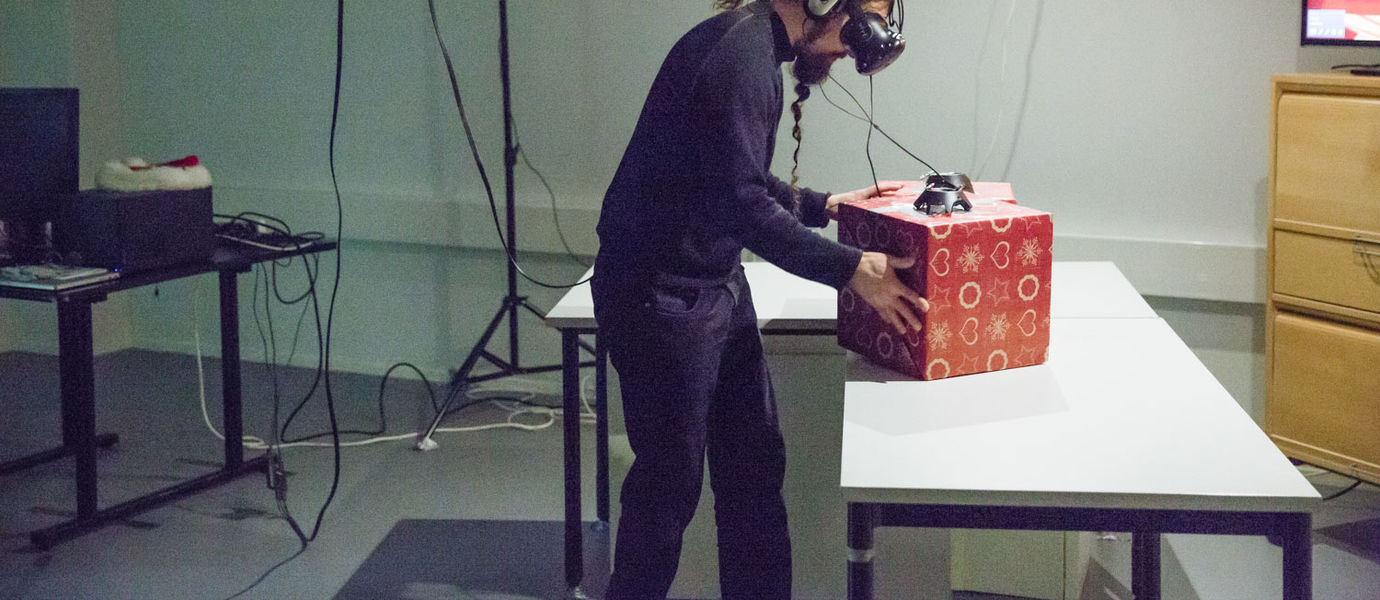 Ein Mann hat Kopfhörer auf und eine VR-Brille. Vor ihm auf dem Tisch stehen zwei rote Pakete. Darauf stehen vermutlich Kameras, die der Mann ansieht. Per Kabel ist er mit ihnen verbunden.