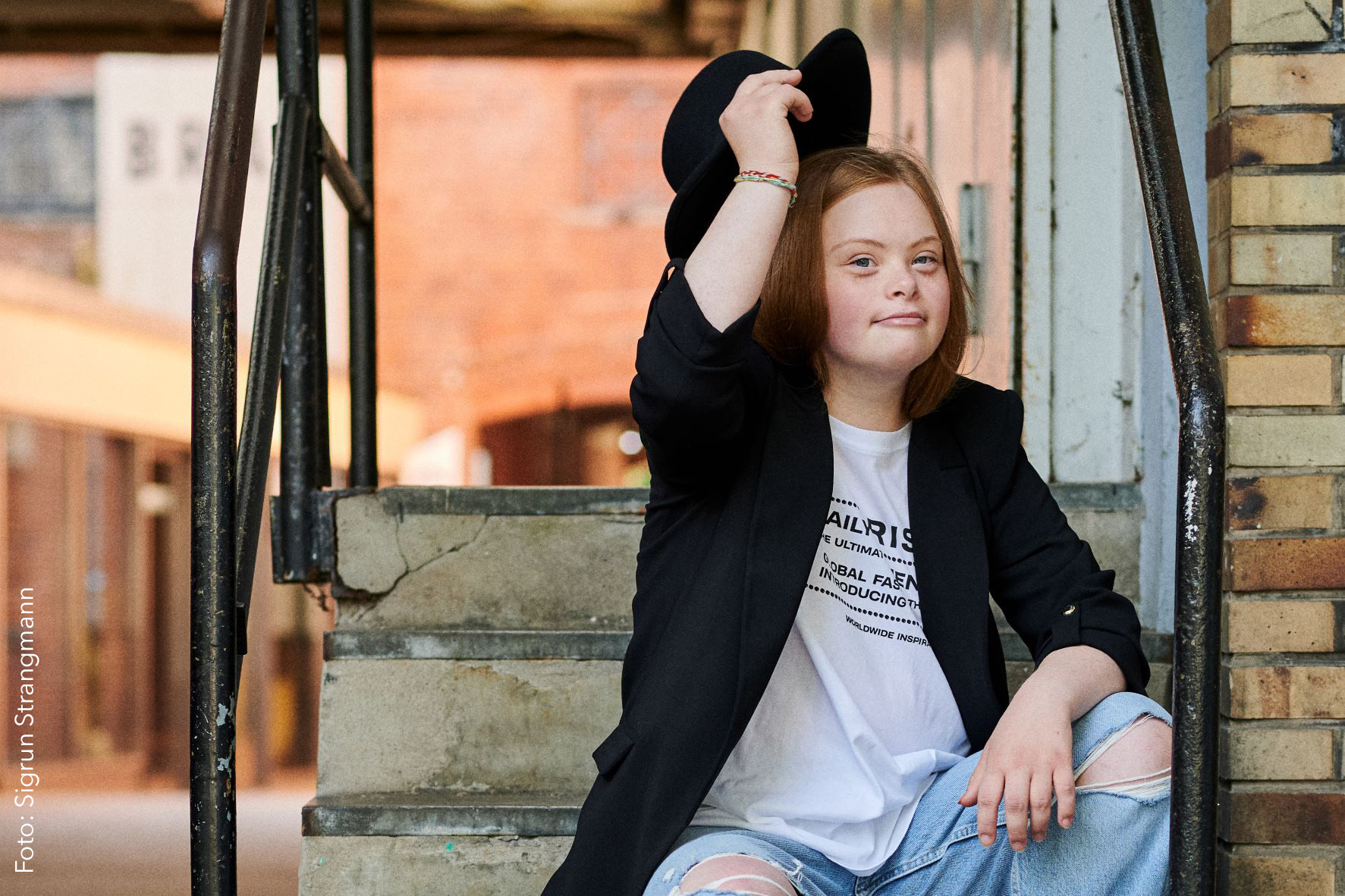 Porträt Amelie Gerdes. Amelie hat lange rotblonde Haare. Sie trägt ein weißes Tshit und einen schwarzen Blazer. Mit ihrer Hand hebt sie gerade einen Hut wie zum Gruße. Sie lächelt und schaut ein bisschen frech. 