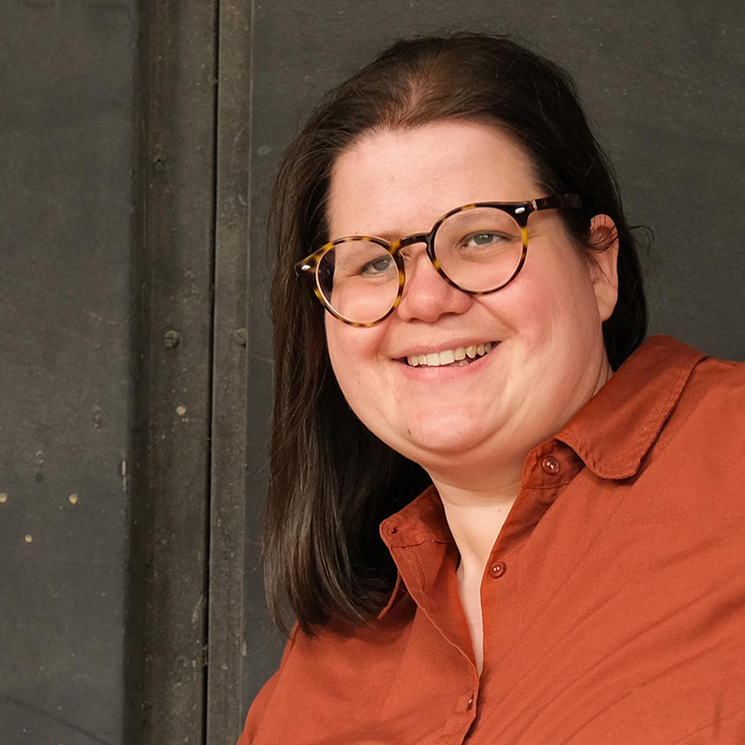 quadratisches Porträt von Ramona in orangener Bluse. Der Hintergrund ist dunkel. Sie trägt eine Brille mit dunklem Rahmen, hat lange schwarze glatte Haate und schaut gerade in die Kamera. Sie hat helle Haut und ein kleines Lächeln auf den Lippen. 