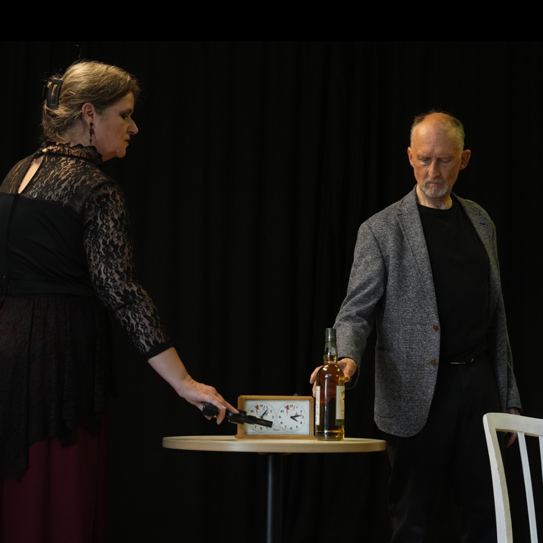 Zwei Erwachsene stehen an einem Tisch mit Schachuhr. Die Frau trägt eine schwarze Spitzenbluse und hält eine Pistole, der Mann greift nach einer Flasche.