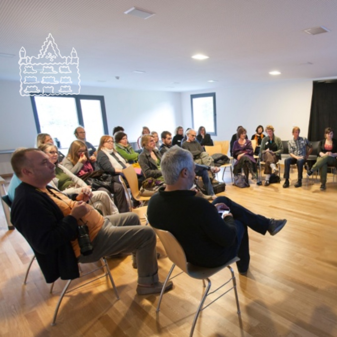 Eine Gruppe von Menschen sitzt in einem Veranstaltungsraum; oben links ist das Logo des Kunsthaus Kannen zu sehen.