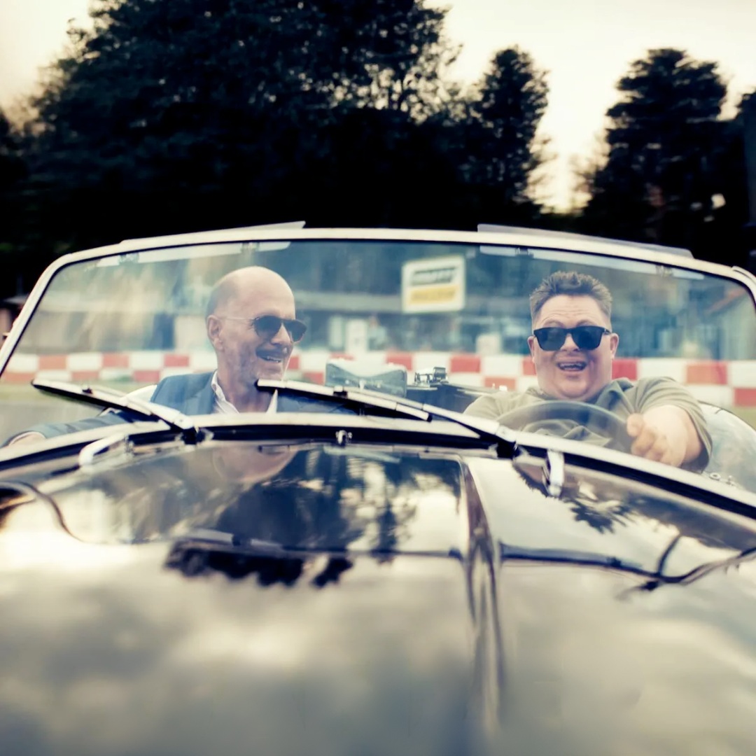 Zwei Männer in einem offenen Oldtimer-Cabrio, beide tragen Sonnenbrillen und lachen. Filmszene aus Ganzer halber Bruder mit Nico Randel und Christoph Maria Herbst.