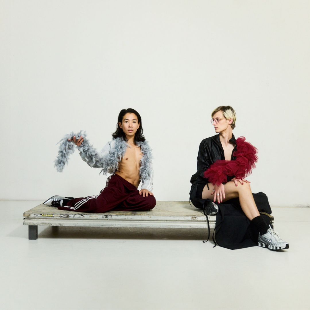 Zwei Personen auf heller Bühne mit weinroten Federboas, entspannt und selbstbewusst, eine auf dem Podest, die andere auf dem Boden.