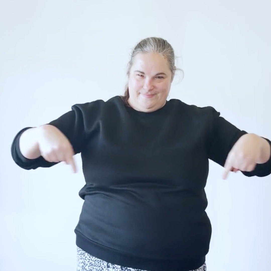 Eine lächelnde Person zeigt mit beiden Fingern nach unten vor hellem Hintergrund.