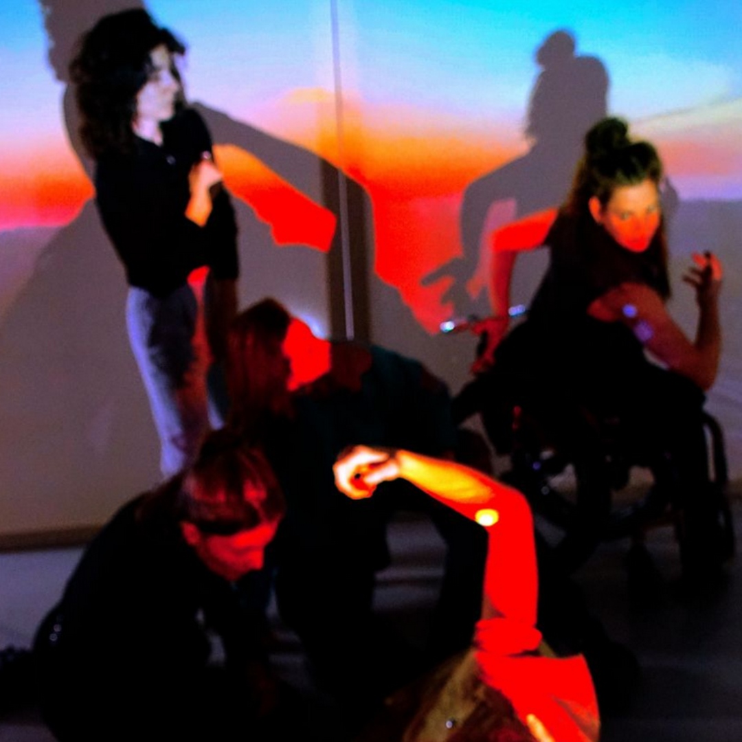 Tanzszene mit mehreren Personen, farbigem Licht und projizierten Schatten an der Wand.