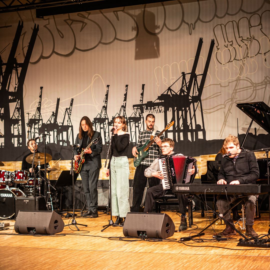 Band mit Sängerin spielt auf einer Bühne vor einer Projektion mit Hafenkränen.