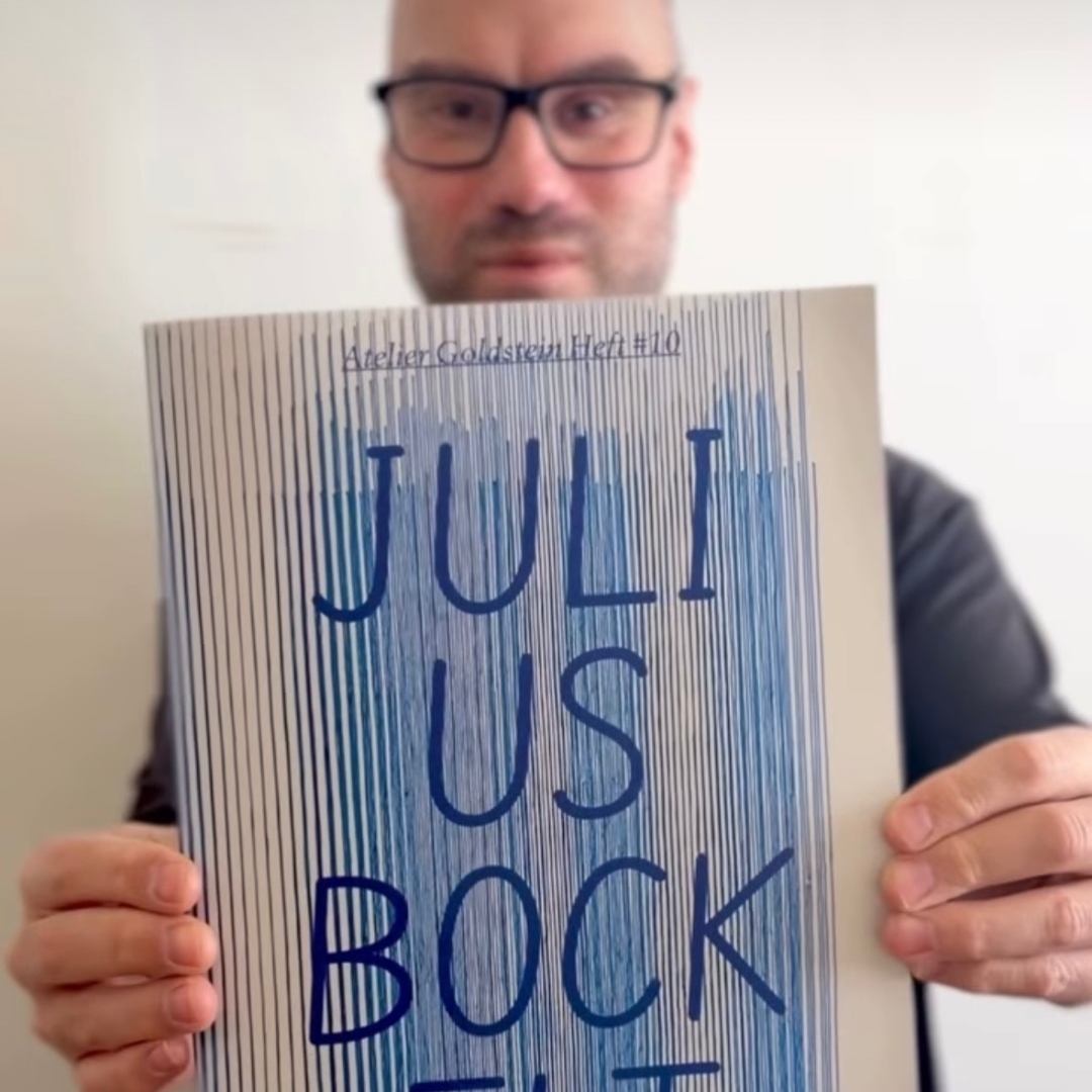 Julius Bockelt hält ein Heft mit blauem Streifenmuster und seinem Namen auf dem Cover vor die Kamera.