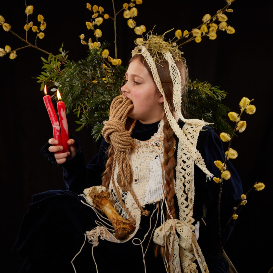 Frau mit Kerzen, Seilen im Mund und Naturmaterialien in einer surrealen Szene.