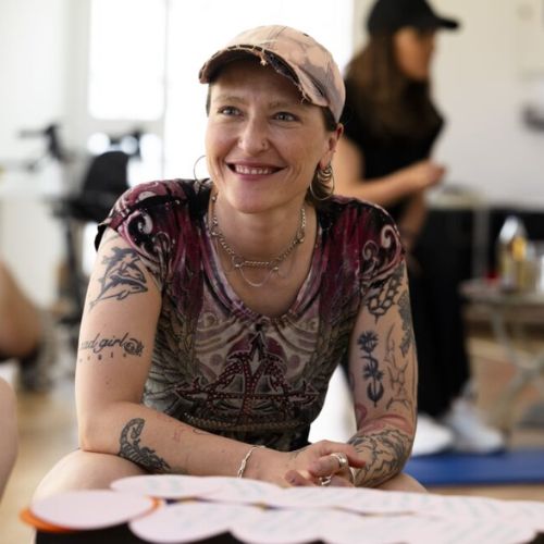 Agnieszka Habraschka Lächelnde Person mit Tattoos und Kappe sitzt an einem Tisch.