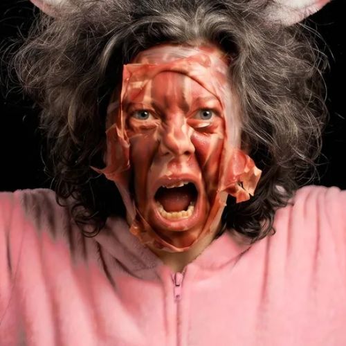Schreiende Frau mit rötlichem, fleischartig wirkendem Material im Gesicht.