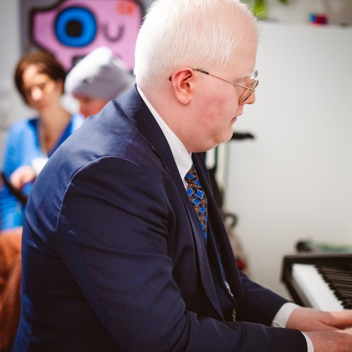 Weißhaariger Mann mit Brille spielt Klavier, im Hintergrund sitzen weitere Personen.