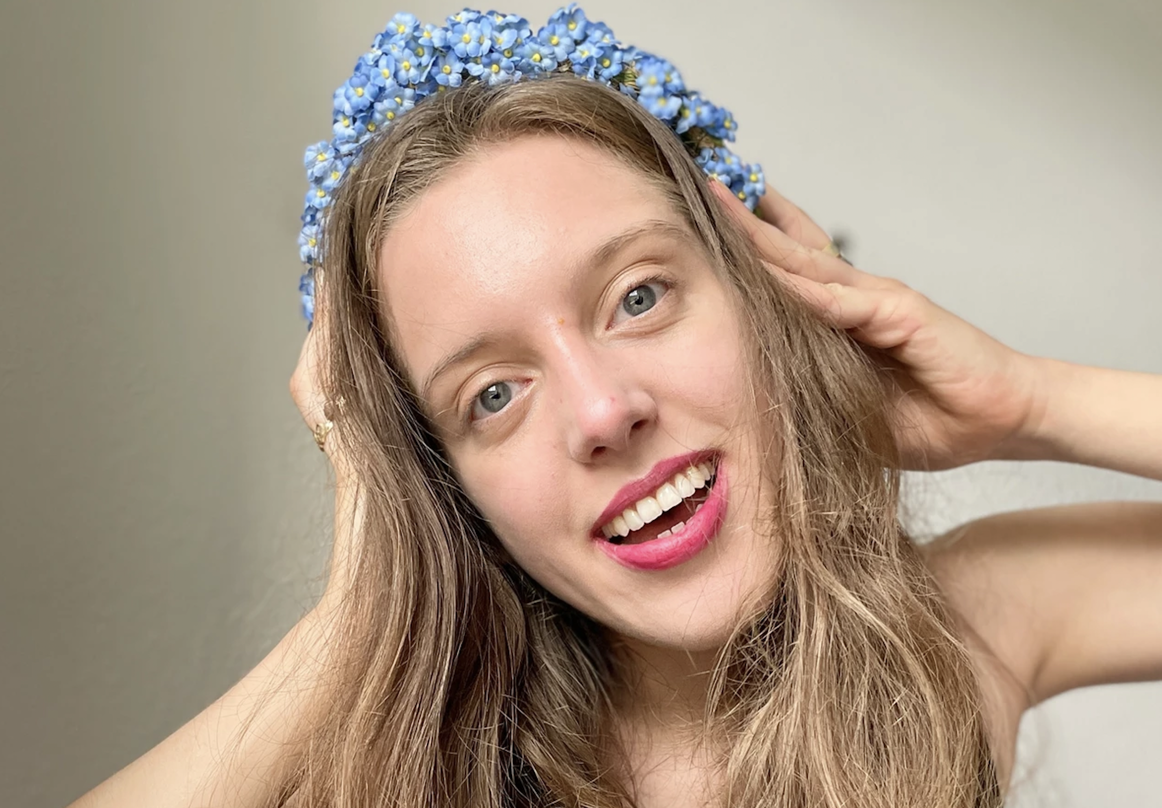 Eine junge, hellhäutige Frau lächelt mit großen, weit aufgerissenen blauen Augen frontal in die Kamera. Lange dunkelblonde Haare umrahmen ihr schmales Gesicht und sie trägt Kopfschmuck aus hellblauen Blümchen.