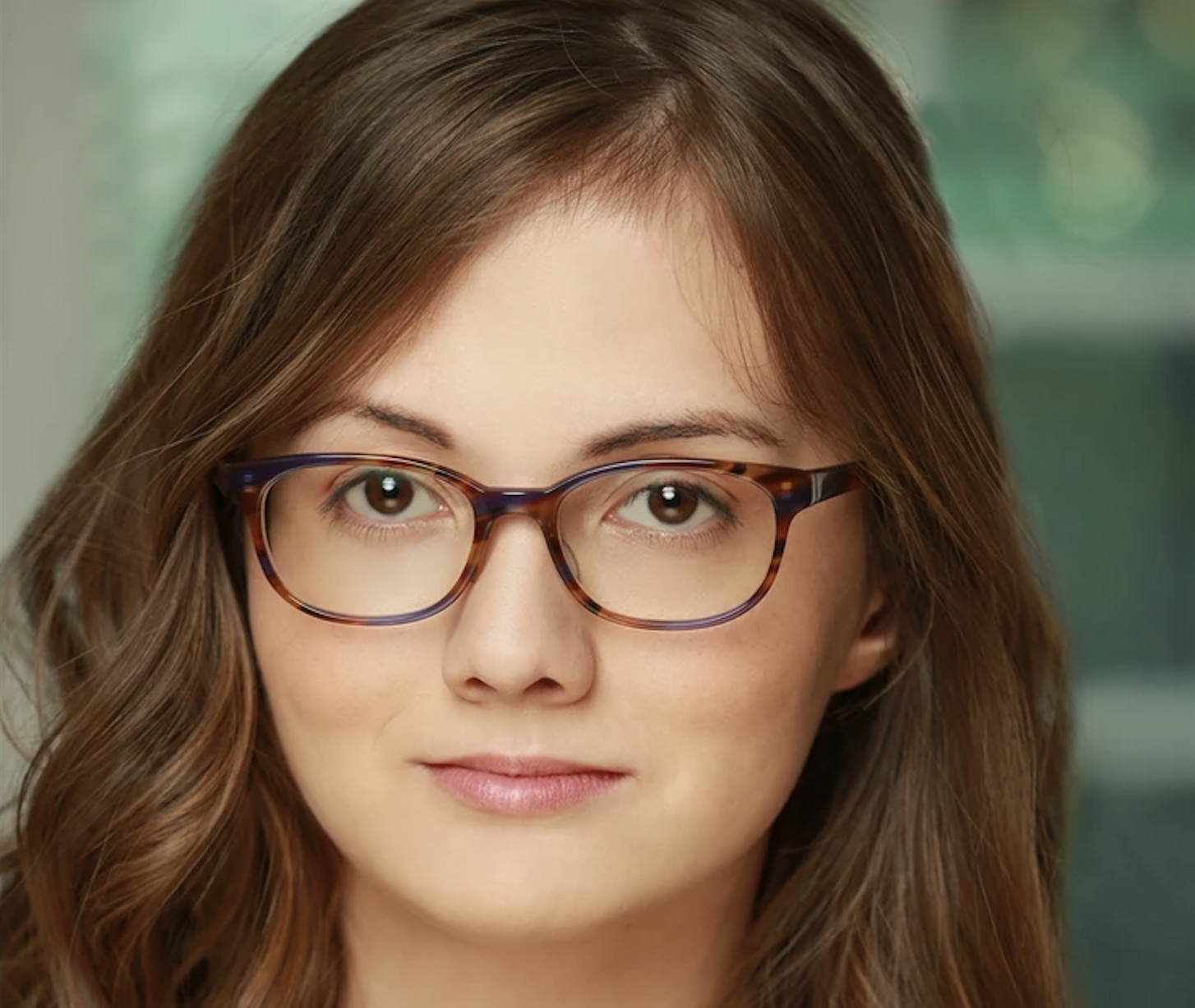 junge Frau mit langen brauen Haaren und Hornbrille lächelt leicht in die Kamera